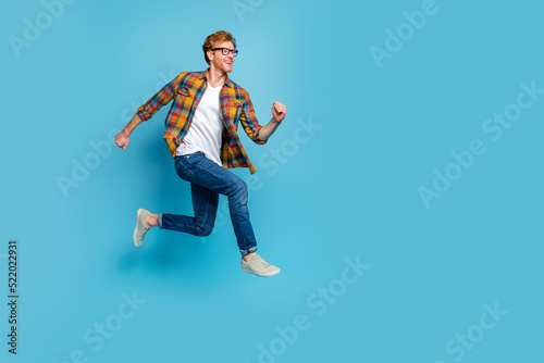 Photo of funky handsome cheerful guy wear checkered shirt with red hairstyle running flying rush isolated on blue color background