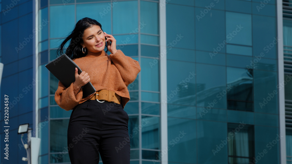 Happy businesswoman stands on background office building speaks on mobile phone answers friendly call communicates with friend using smartphone spends free time outdoors holding folder with documents