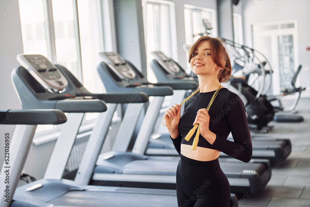 Fototapeta premium Standing with jump rope and smiling. Young beautiful woman in sportive clothes have fitness day in the gym