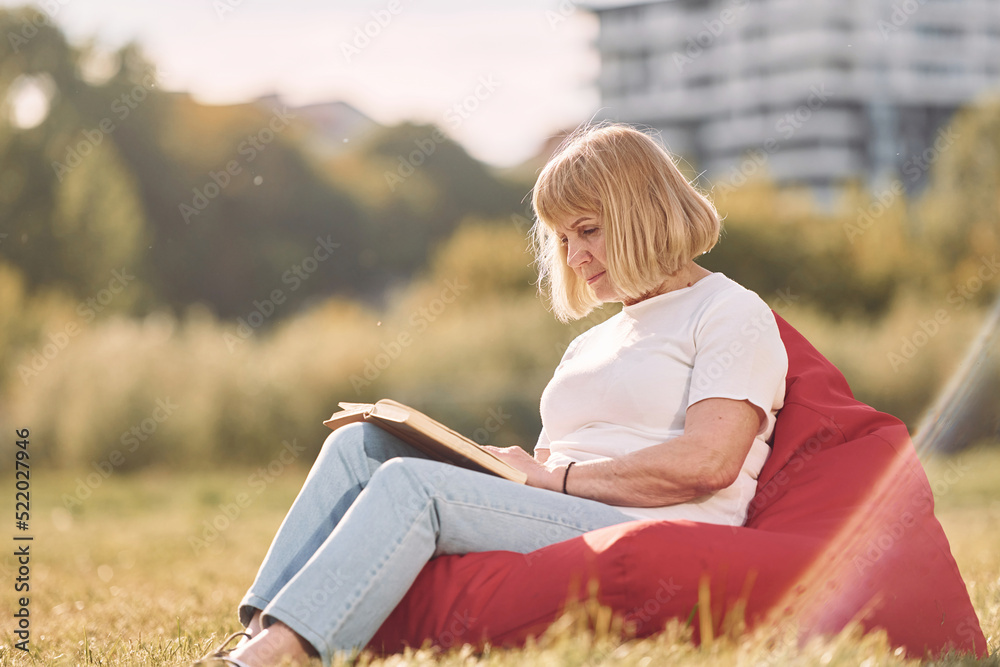 Sitting on the armchair and reading the book. Senior woman having nice weekend outdoors on the field at sunny day Sitting on the armchair and reading the book. Senior woman having nice weekend outdoors on the field at sunny day