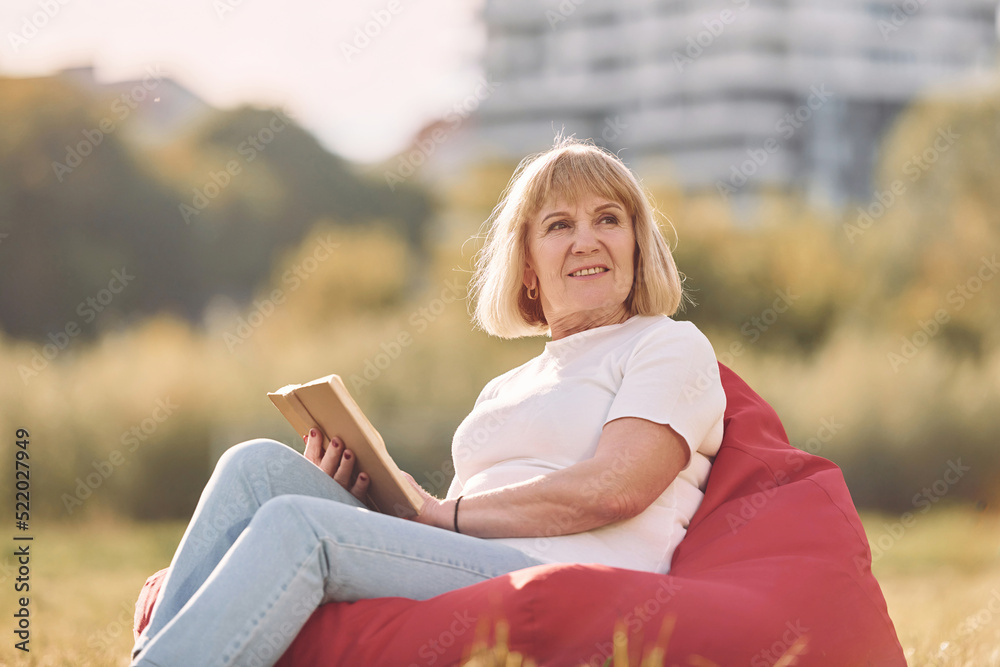 Sitting on the armchair and reading the book. Senior woman having nice weekend outdoors on the field at sunny day Sitting on the armchair and reading the book. Senior woman having nice weekend outdoors on the field at sunny day