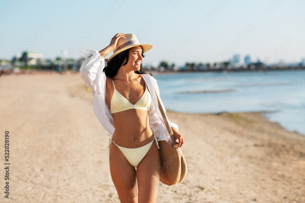 people, summer holidays and leisure concept - happy young woman in bikini swimsuit, white shirt and straw hat with bag walking along beach people, summer holidays and leisure concept - happy young woman in bikini swimsuit, white shirt and straw hat with bag walking along beach