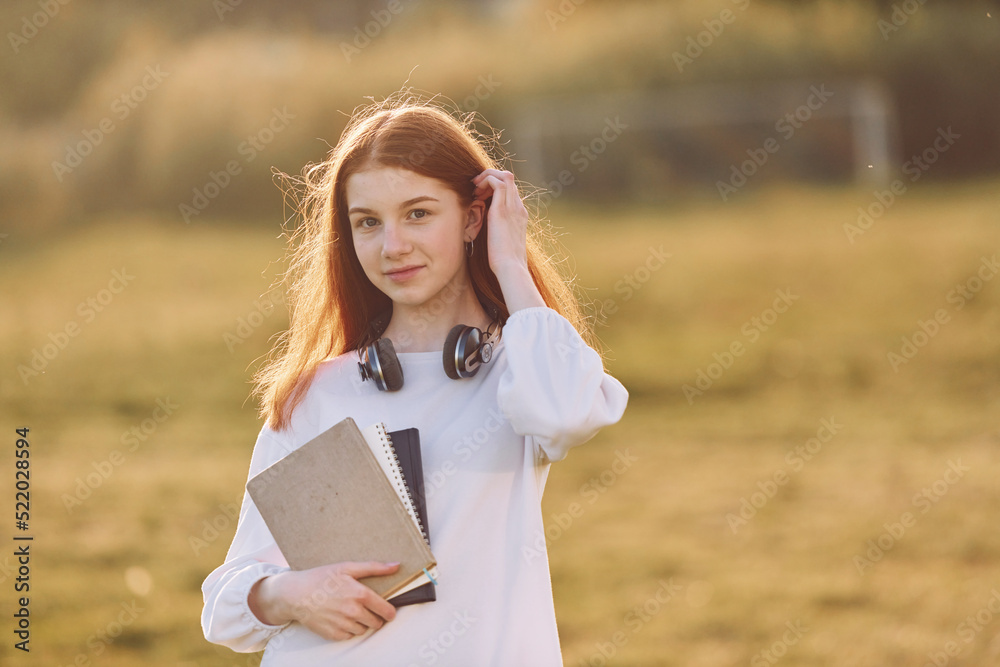 Naklejka premium Student with notepad and with headphones. Young girl is on the field at sunny daytime having nice weekend