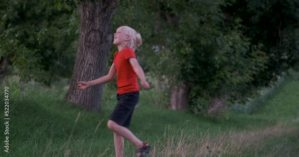 Child has fun, spinning and dancing nature. Blond hair boy whirls on lawn in forest. Boy in red t-shirt in forest Child has fun, spinning and dancing nature. Blond hair boy whirls on lawn in forest. Boy in red t-shirt in forest