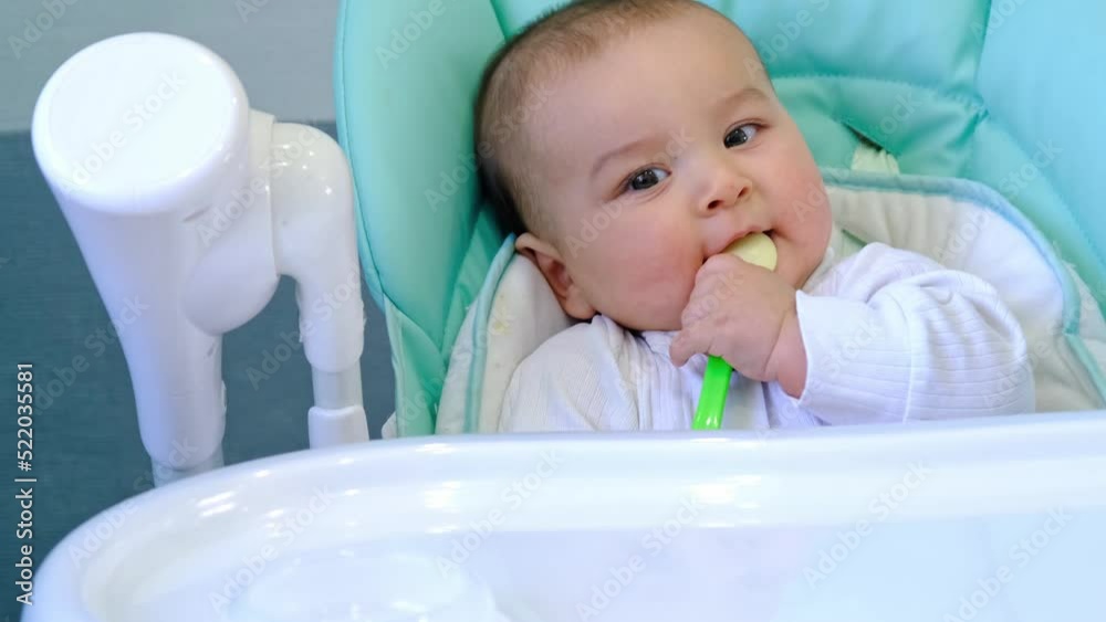 A hungry baby is gnawing on a plastic spoon at the table on a high chair. Teething, whims, itchy gums, introduction of complementary foods A hungry baby is gnawing on a plastic spoon at the table on a high chair. Teething, whims, itchy gums, introduction of complementary foods