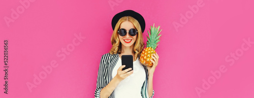 Summer portrait of happy smiling woman with smartphone and pineapple wearing black hat, sunglasses on pink background