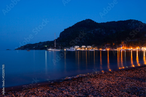 Night view of the embankment and promenade in the light of street lights and lighting of restaurants. small town surrounded by mountains in evening. Light reflections on sea water surface. Night beach