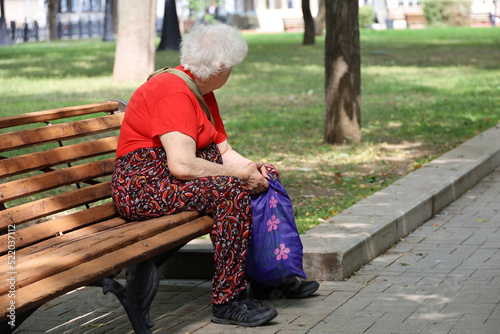 Fototapeta Naklejka Na Ścianę i Meble -  Elderly gray-haired woman sitting with shopping bag on a bench in a park. Leisure in summer city, life in retirement