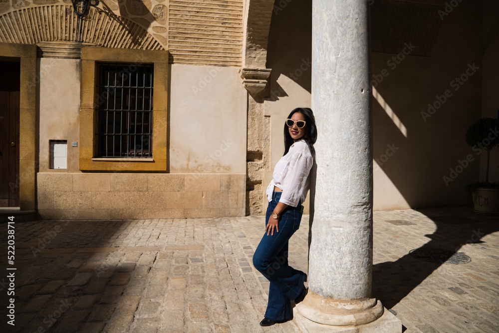 young, beautiful, brunette south american woman is travelling in europe and is leaning on a white marble column in a typical street of an old european city. Travel and holiday concept.