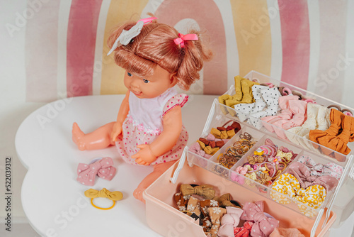 Baby doll with blonde hair sitting on white table and organizer with hair bows in Children's room. Nursery Organizing and Storage Ideas. Baby Girl accessory. Hairstyles for girls with bows.