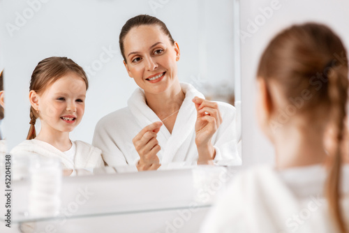 beauty, hygiene, morning and people concept - happy smiling mother and little daughter with dental floss cleaning teeth and looking to mirror at bathroom