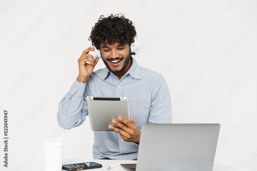 Young indian smiling man sitting with laptop looking on tablet Young indian smiling man sitting with laptop looking on tablet