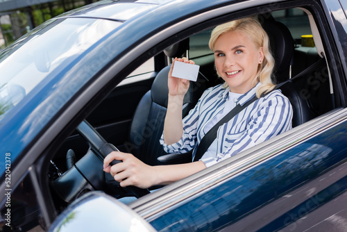 Smiling blonde woman holding driver license on driver seat in car