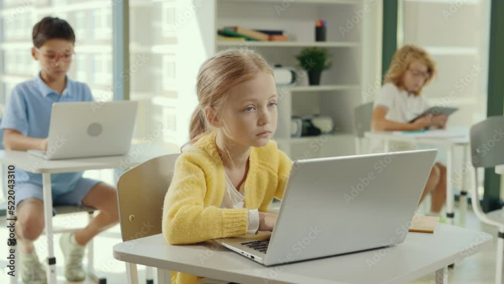 School girl using the computer sitting in the class during computer science lesson and other pupils sitting and using computers or tablets on the background. People and technology concept.