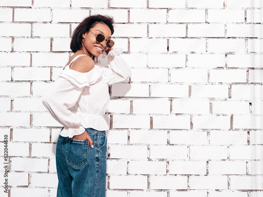 Portrait of young beautiful black woman. Smiling model dressed in summer jeans clothes. Sexy carefree female posing near white brick wall in studio. Tanned and cheerful. In sunglasses Portrait of young beautiful black woman. Smiling model dressed in summer jeans clothes. Sexy carefree female posing near white brick wall in studio. Tanned and cheerful. In sunglasses