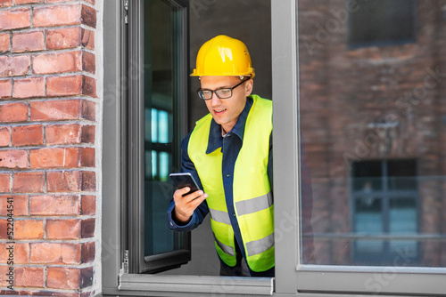 construction business and building concept - smiling male builder in helmet and safety west with smartphone at open window