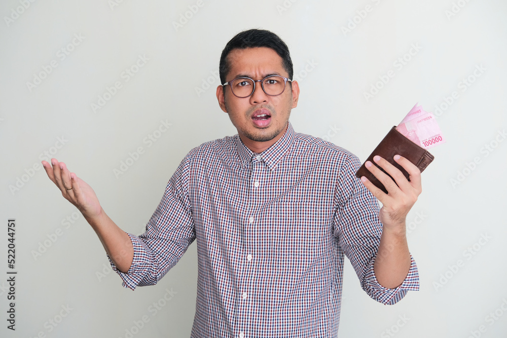 Adult Asian man holding wallet with money inside and showing confused expression Adult Asian man holding wallet with money inside and showing confused expression