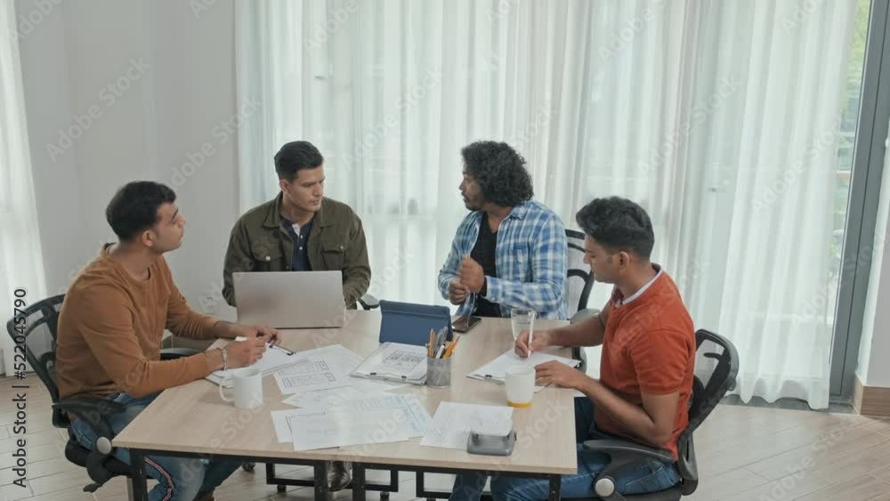 From above slowmo of young male Indian entrepreneurs sitting around office desk having discussion about business project From above slowmo of young male Indian entrepreneurs sitting around office desk having discussion about business project