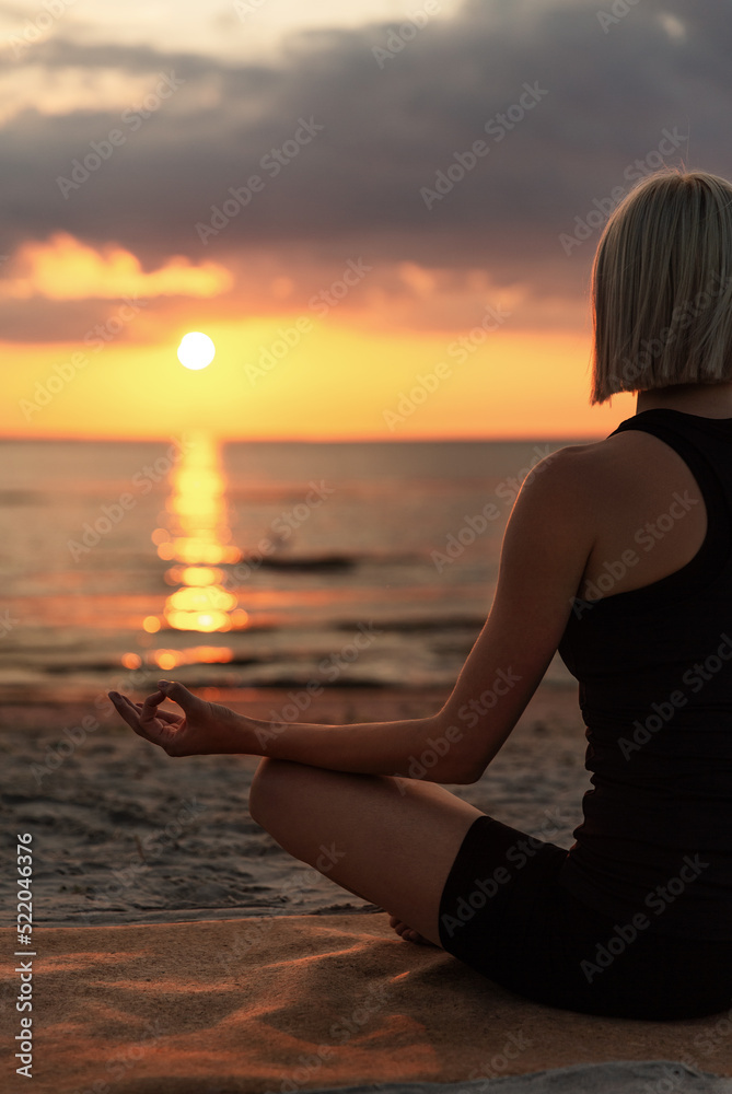 yoga, mindfulness and meditation concept - woman meditating in lotus pose on beach over sunset