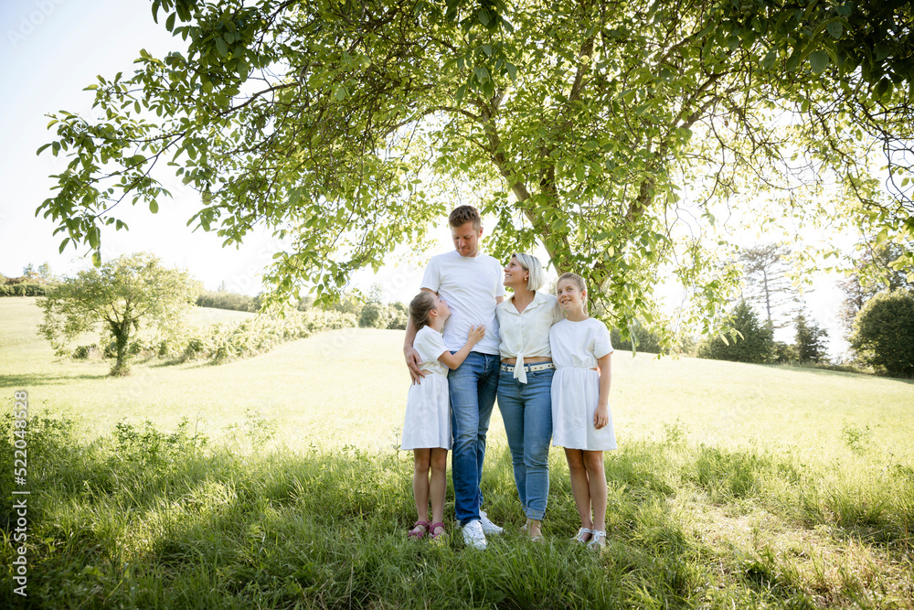 beautiful family with two girls is standing in meadow near walnut tree and has light outfit on and jeans and is happy