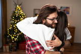 A man with glasses and beard hugs girlfriend, whom he has not seen for long time. Brunette with long hair wearing red plaid shirt. Behind them shimmers a Christmas tree.