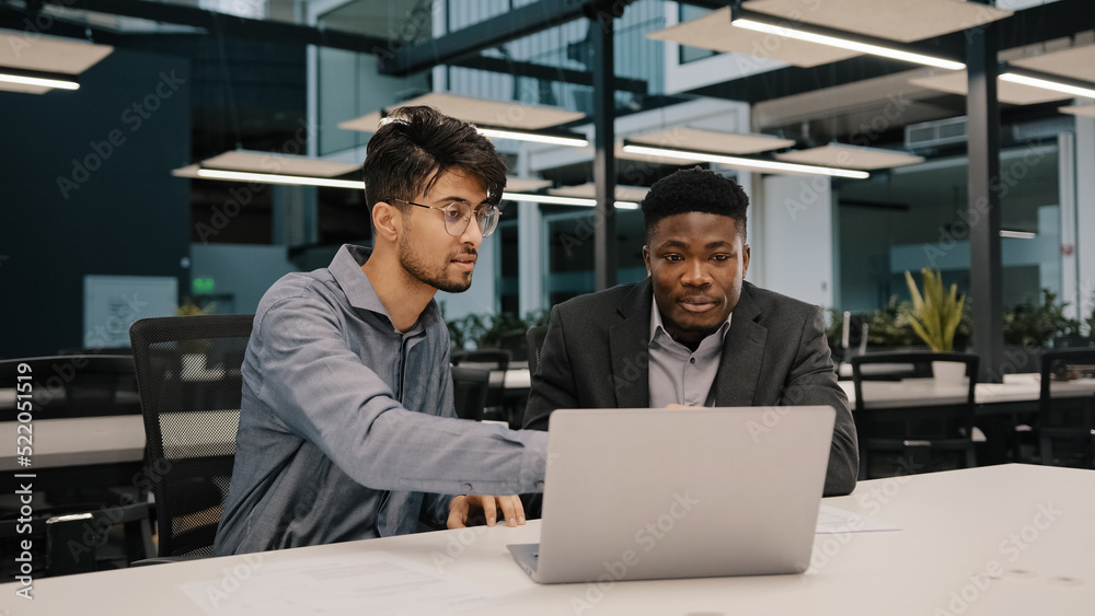 Two multicultural professional male co-workers men colleagues sit at office use laptop discuss internet project hold negotiation choosing business profitable contract cooperation teamwork partnership
