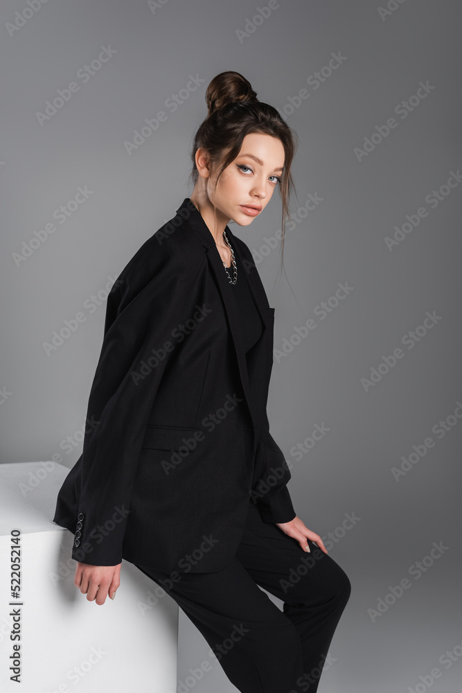 young and trendy woman in black blazer looking at camera near white cube isolated on grey young and trendy woman in black blazer looking at camera near white cube isolated on grey