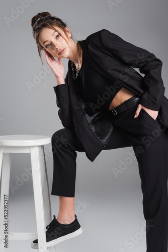 pretty woman in black clothes posing with hand in pocket near white stool on grey background