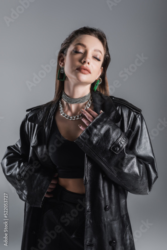 sensual woman in leather clothes and silver necklaces posing with closed eyes isolated on grey