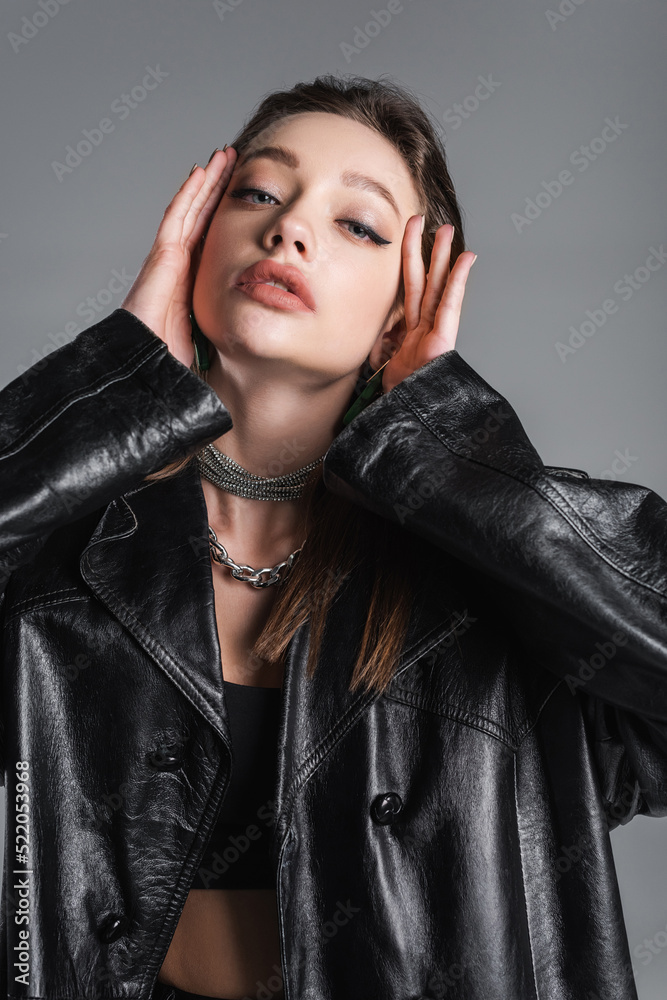 brunette woman in black leather coat and makeup touching face isolated on grey brunette woman in black leather coat and makeup touching face isolated on grey