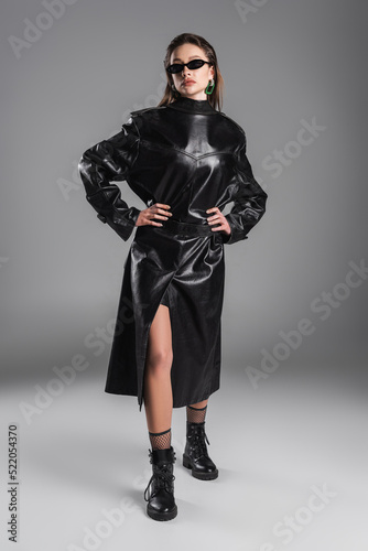 full length of woman wearing black leather coat back to front and posing with hands on hips on grey background