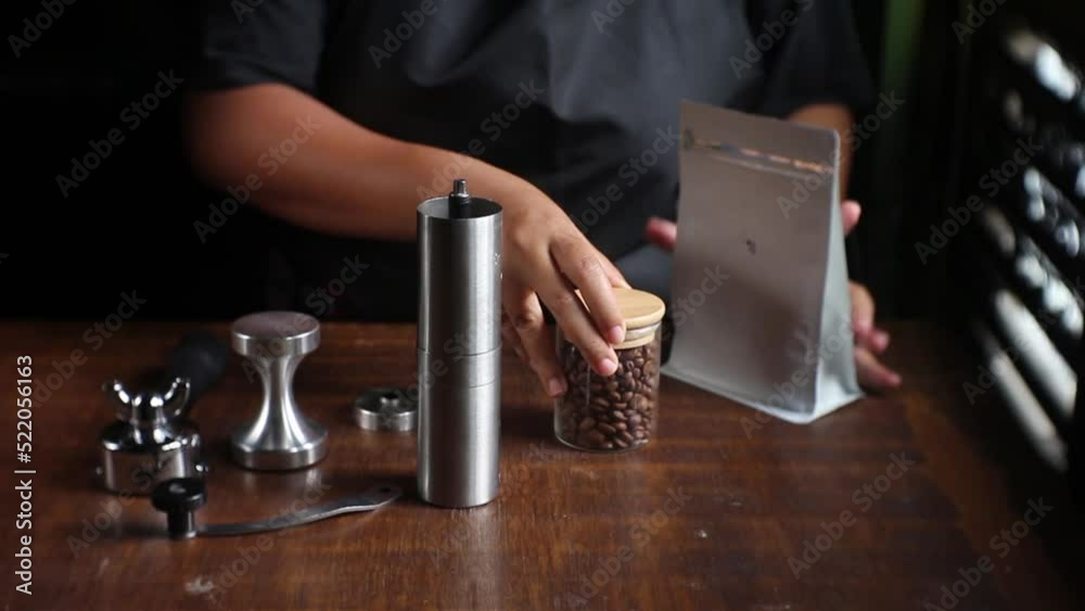 Dark roasted bean poured by home barista into metal hand grinder on a brown wooden bar table. Cafe activities for making V60 coffee brew