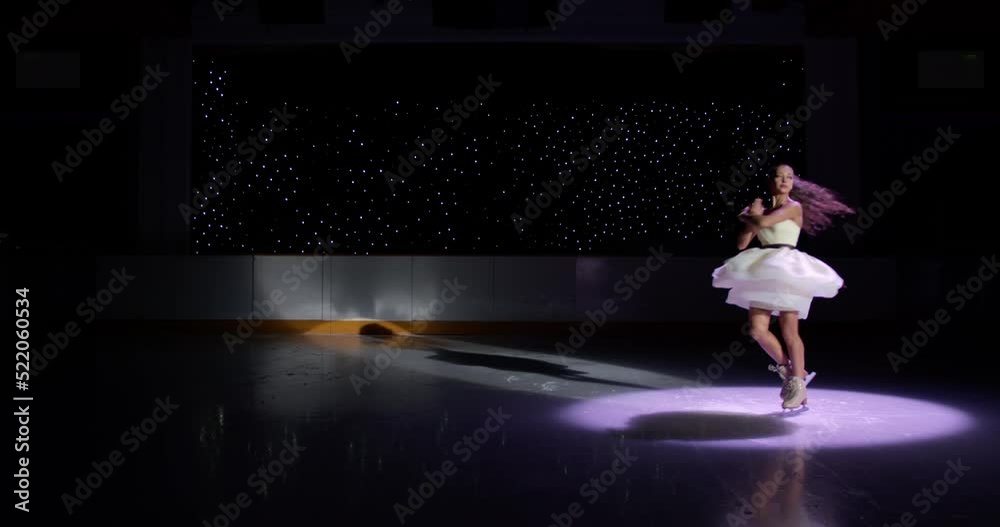 Ice skater girl in white dress spins on dark ice rink in spotlight, figure skating performance