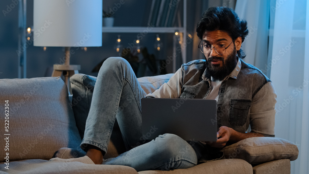 Businessman guy Arab Indian man bearded male user wearing glasses sitting on sofa at night late time with laptop reading bad news online trouble app job problems pondering solution looking at screen