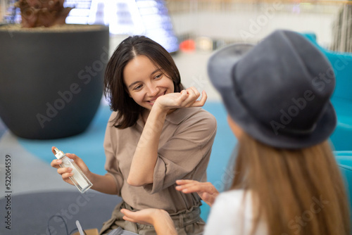 Happy young woman smelling her perfumed hand in shopping mall. Smiling female friends sitting and discussing perfume indoors. Beauty and female friendship concept