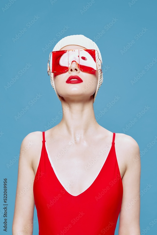 A bright red-lipped lady in a red swimsuit glasses swimming cap raises her head up posing isolated on a blue background in the studio