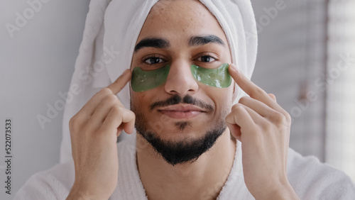 Front view portrait male face smiling indian arabian man wearing bath towel head glue sticky hydrogel eye-patches cosmetic procedures skin care guy looking at camera with collagen patches under eyes