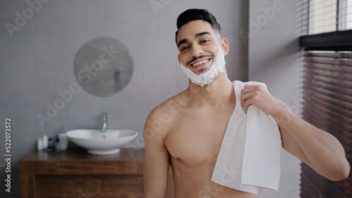 Close up male portrait in bathroom hispanic indian arabian naked man bearded guy posing smiling with white soapy foam on beard shaving gel holding bath towel looking at camera getting ready to shave
