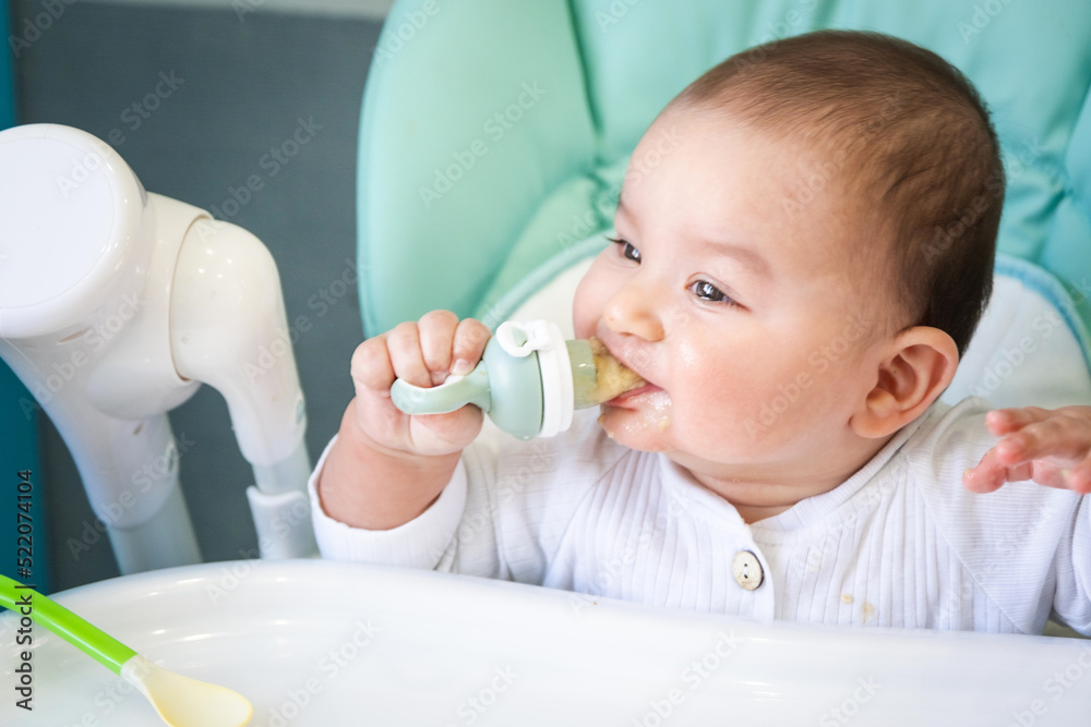 A happy and contented baby eats banana puree from a nipple. Introduction of complementary foods, healthy baby food, feeding the baby at the table A happy and contented baby eats banana puree from a nipple. Introduction of complementary foods, healthy baby food, feeding the baby at the table