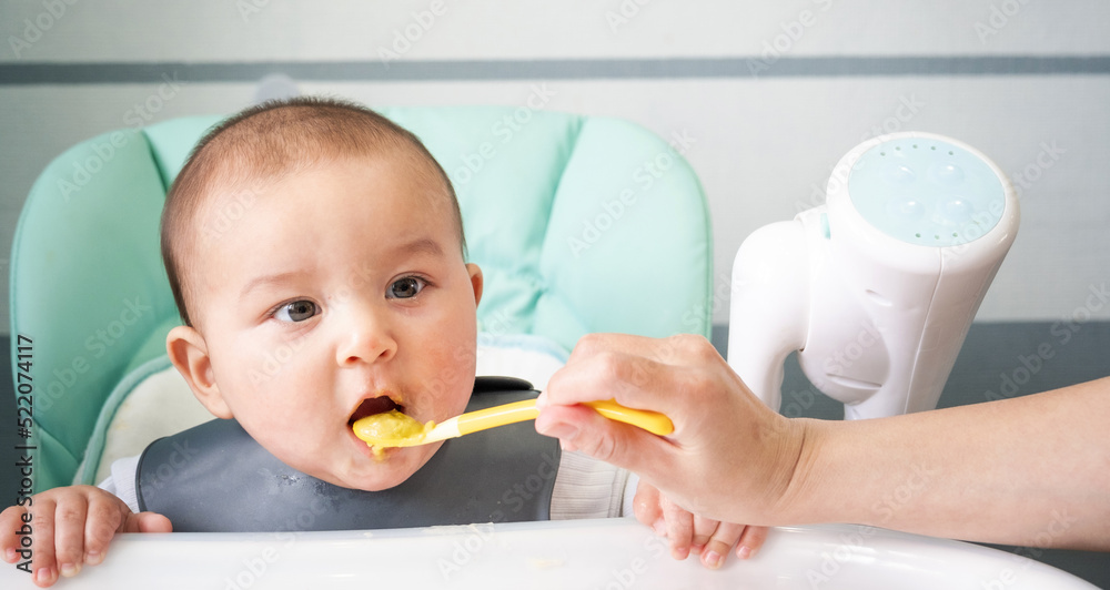 Mom feeds the baby with a spoon of vegetable puree at the children's feeding table. Baby's appetite, healthy nutrition, introduction of complementary foods. Copyspace, mock up Mom feeds the baby with a spoon of vegetable puree at the children's feeding table. Baby's appetite, healthy nutrition, introduction of complementary foods. Copyspace, mock up