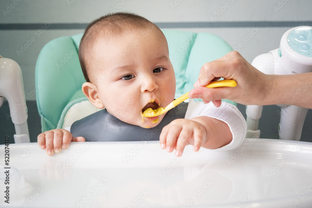 Mom feeds the baby with a spoon of vegetable puree at the children's feeding table. Baby's appetite, healthy nutrition, introduction of complementary foods. Copyspace, mock up Mom feeds the baby with a spoon of vegetable puree at the children's feeding table. Baby's appetite, healthy nutrition, introduction of complementary foods. Copyspace, mock up