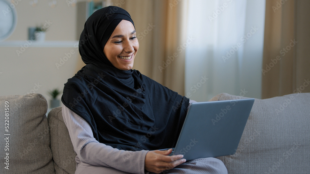 Happy cheerful islamic young girl making video call sit in living room enjoy pleasant online conversation carefree muslim smiling woman using laptop for remote communication modern technology concept Happy cheerful islamic young girl making video call sit in living room enjoy pleasant online conversation carefree muslim smiling woman using laptop for remote communication modern technology concept