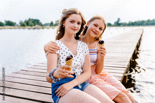 Two cheerfull teenager girls sitting on wooden bridge by the lake. Outdoor travel vacation healthy lifestyle vegan food picnic on beach hungry children with snacks. photo