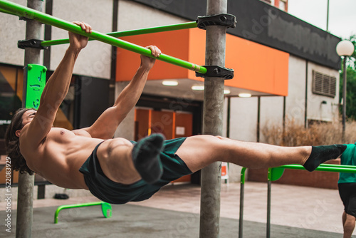 Front view of a muscular shirtless caucasian man exercising on a calisthenics outdoor bars.
