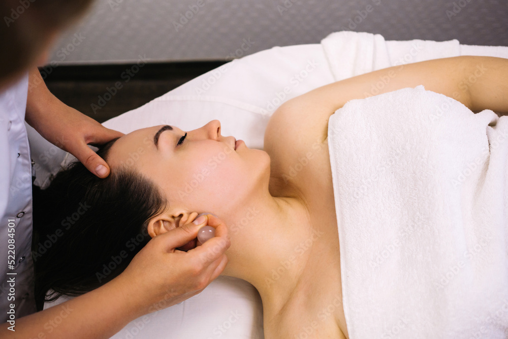 Woman receiving facial massage with gua sha tool in beauty salon. Beauty and skincare concept with a beautiful woman. Middle aged female using guasha. Massage for facial lifting Woman receiving facial massage with gua sha tool in beauty salon. Beauty and skincare concept with a beautiful woman. Middle aged female using guasha. Massage for facial lifting