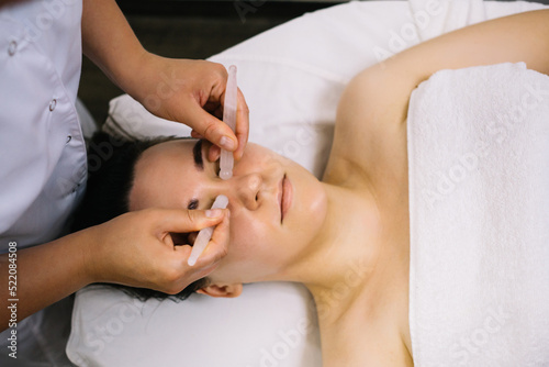 Woman receiving facial massage with gua sha tool in beauty salon. Beauty and skincare concept with a beautiful woman. Middle aged female using guasha. Massage for facial lifting