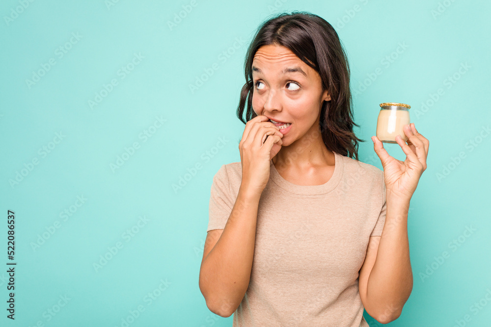 Young hispanic woman holding yogurt isolated on blue background relaxed thinking about something looking at a copy space. Young hispanic woman holding yogurt isolated on blue background relaxed thinking about something looking at a copy space.