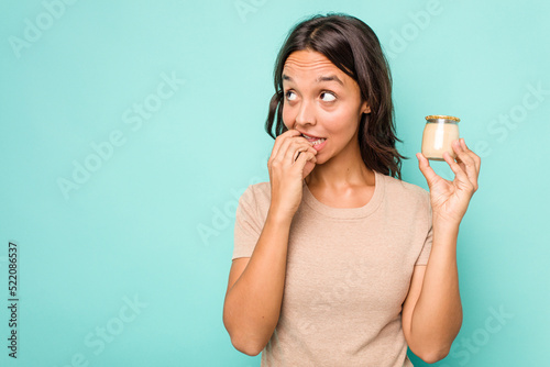 Young hispanic woman holding yogurt isolated on blue background relaxed thinking about something looking at a copy space.