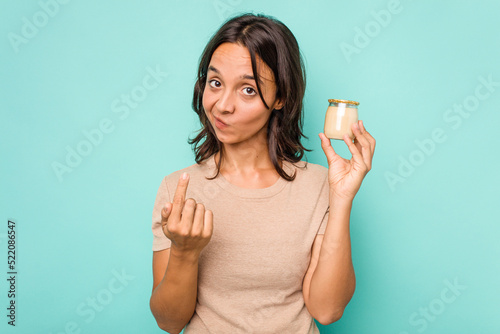 Young hispanic woman holding yogurt isolated on blue background pointing with finger at you as if inviting come closer.