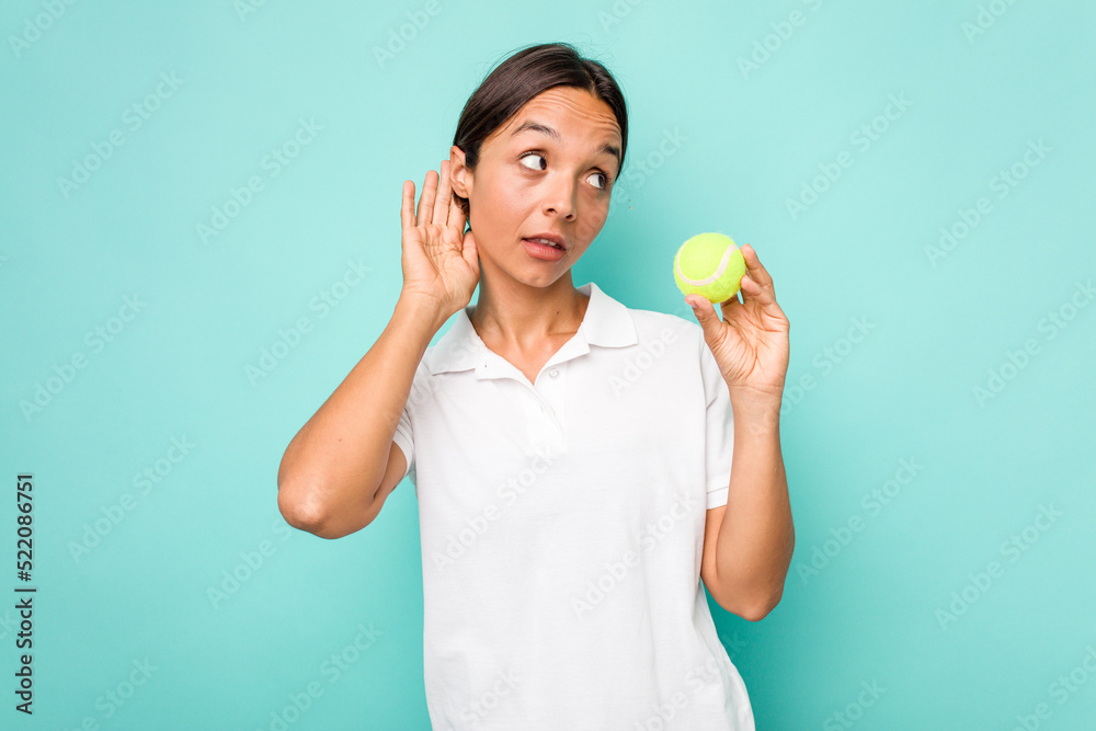 Young hispanic physiotherapy holding a tennis ball isolated on blue background trying to listening a gossip. Young hispanic physiotherapy holding a tennis ball isolated on blue background trying to listening a gossip.